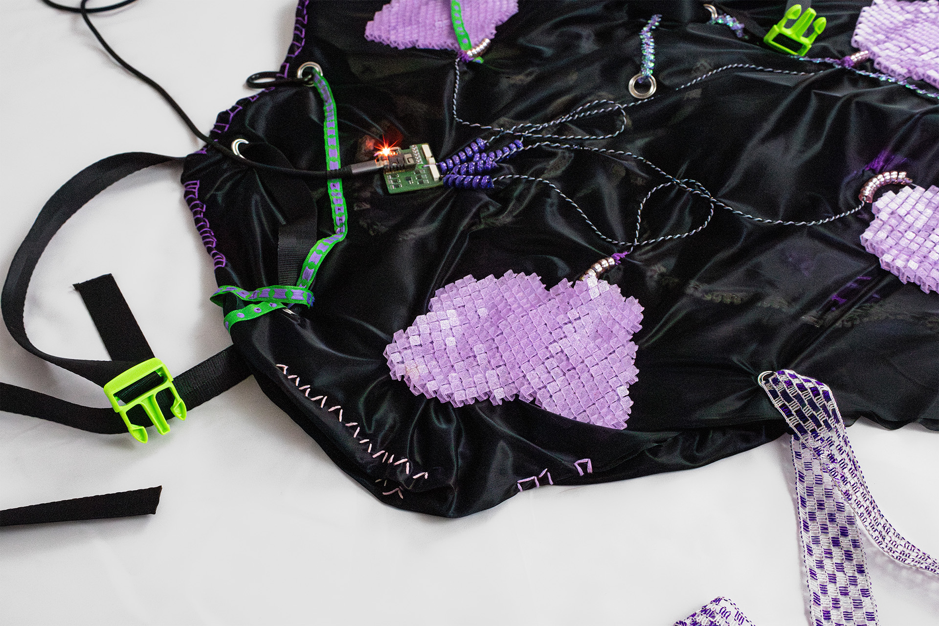 On the photo, a Tactile Patch is shown. It takes an indeterminate shape, but can be most easily describes as a small blanket. To attach it to a body, straps like those of a backpack are attached at various points. Four violet 3D printed fabric panels are sewn onto a black shiny fabric, and they are connected to a small Printed Circuit Board via cables. Every 3D printed panel holds a vibration motor within that is not visible but can be felt buzzing.