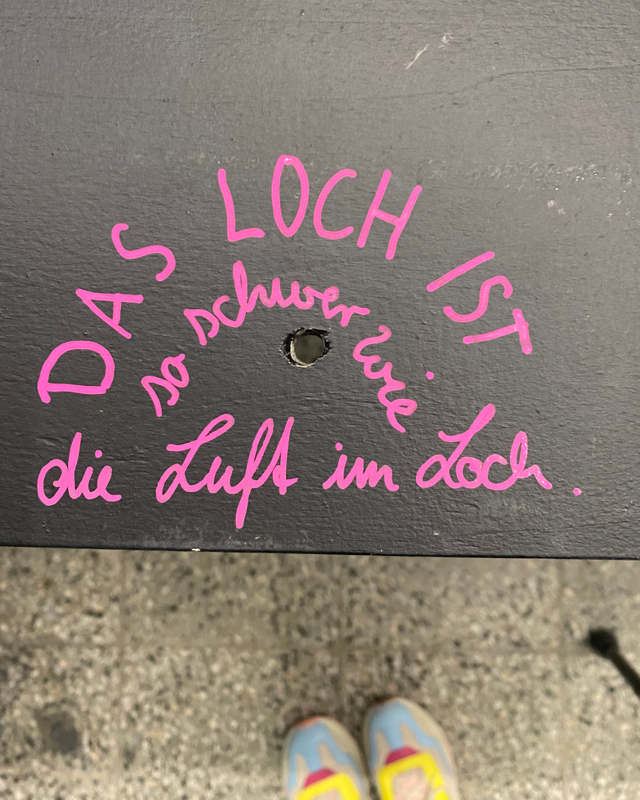 Photograph of a short hand written tactile description in pink that wraps around a small hole drilled through a dark table-like display. The descriptions reads: Das Loch ist so schwer wie die Luft im Loch. In English this translates to: The hole is as heavy as the air within the hole.