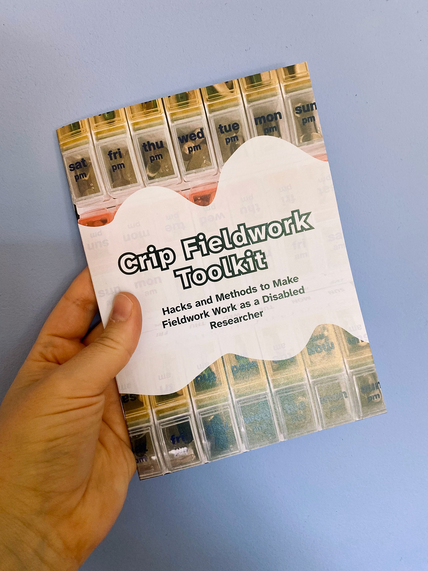A white person's hand is holding the Crip Fieldwork Toolkit booklet into the camera. On the cover, the title reads: Crip Fieldwork Toolkit. Hacks and Methods to Make Fieldwork Work as a Disabled Researcher. This text is displayed atop a curved white banner, that sits playfully stop a vivid photgraph of a pillbox. The pills form a mesmerizing pattern.