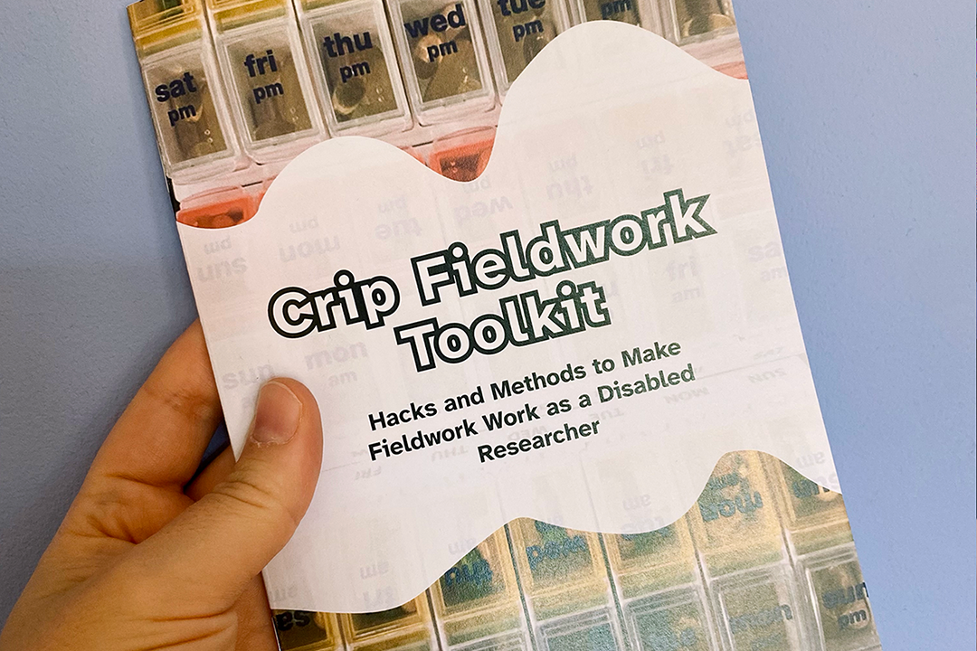 A white person's hand is holding the Crip Fieldwork Toolkit booklet into the camera. On the cover, the title reads: Crip Fieldwork Toolkit. Hacks and Methods to Make Fieldwork Work as a Disabled Researcher. This text is displayed atop a curved white banner, that sits playfully stop a vivid photgraph of a pillbox.