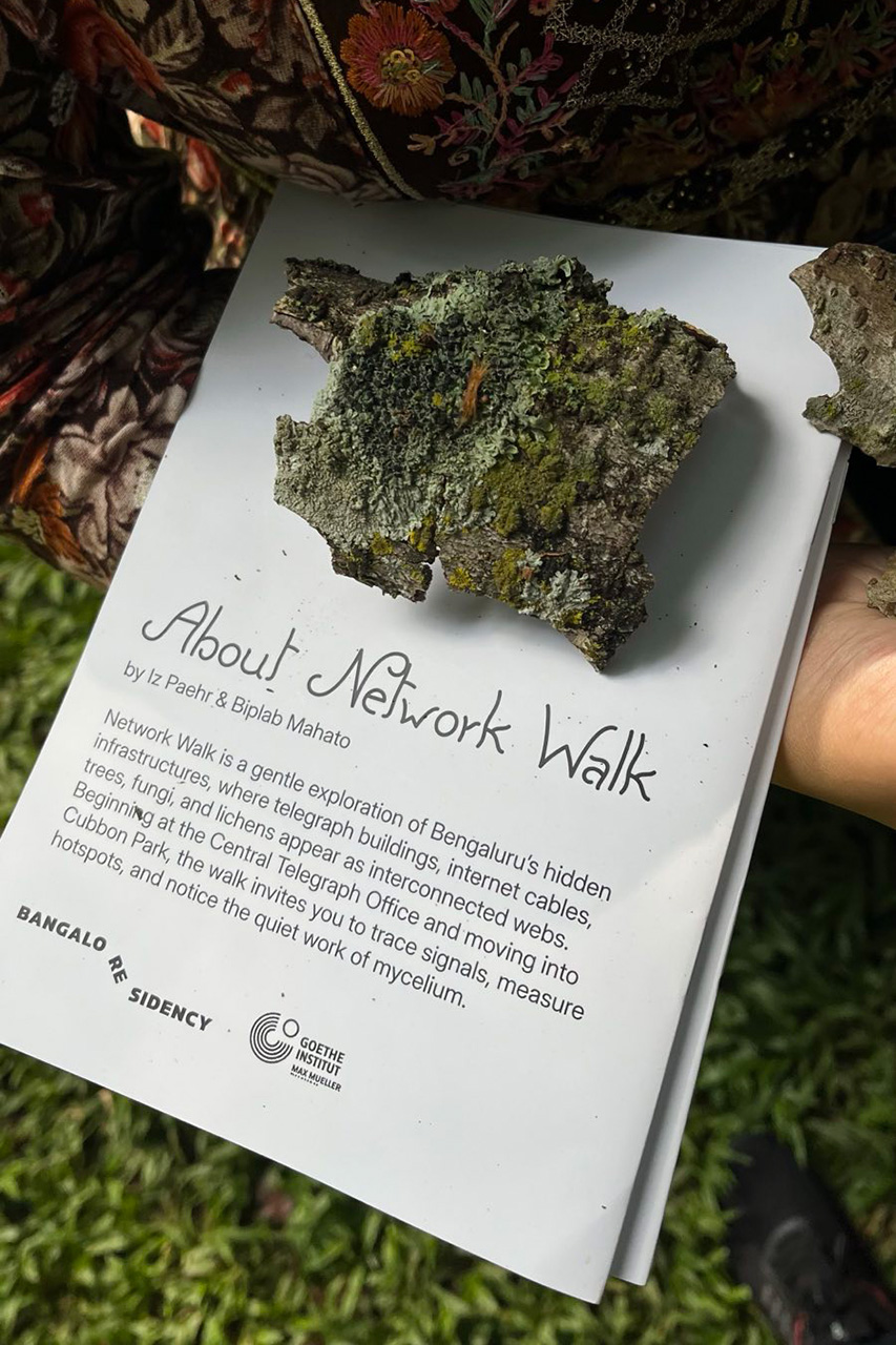 A piece of park with lichens and moss balances atop a network walk booklet held by a person in a beautifully embroidered shirt. The text printed on the booklet reads: About Network Walk, with a short abstract about the walk underneath.