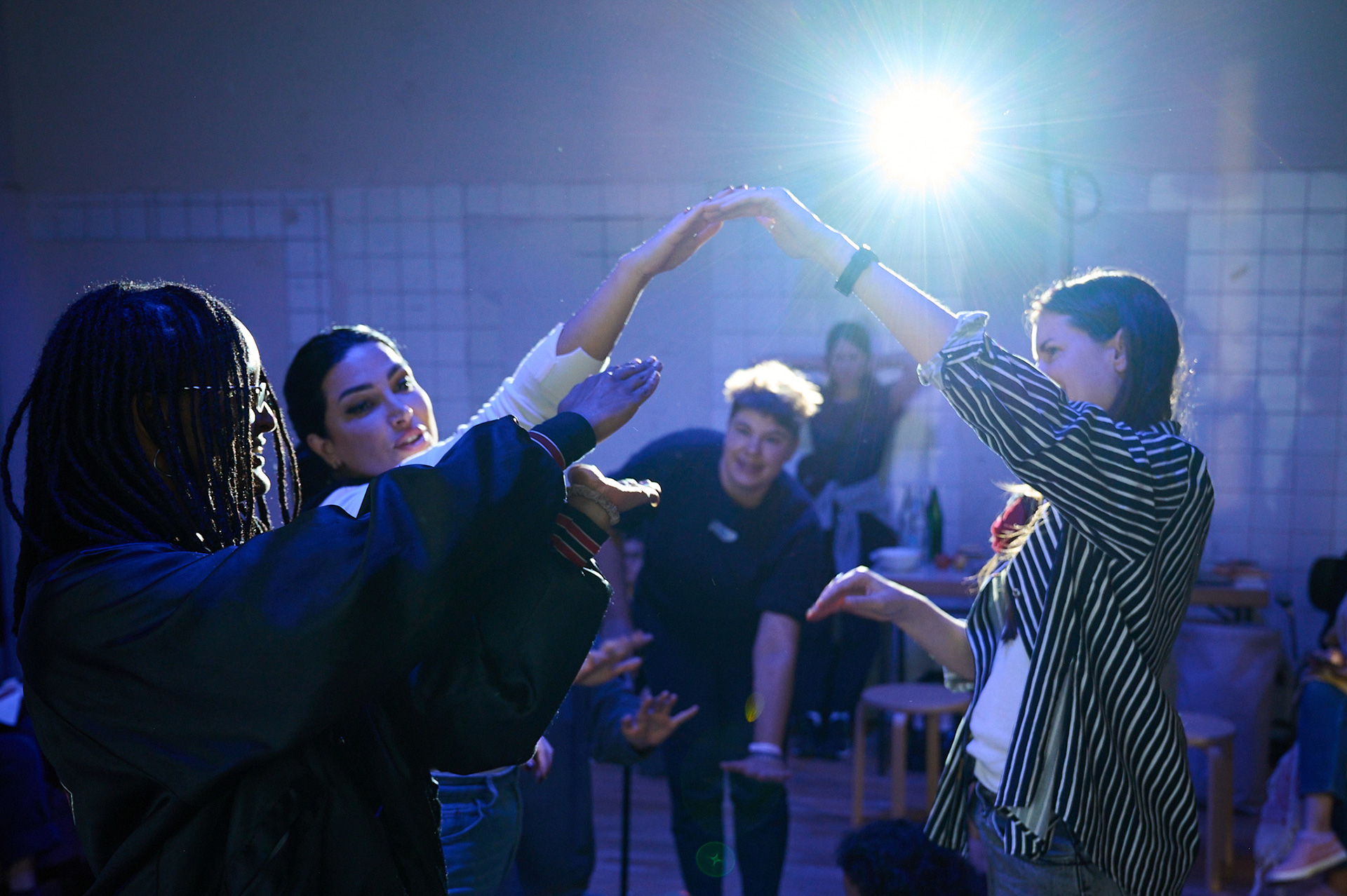 A projector in a dark studio spaces shines light on the following scene: Three women are raising their hands and perform movements as though they are touching an invisible object. In the backrgound, Iz is entering the scene. They are holding one of their hands stretched out towards the ground, while the other hand holds their walking stick. The atmosphere is playful.