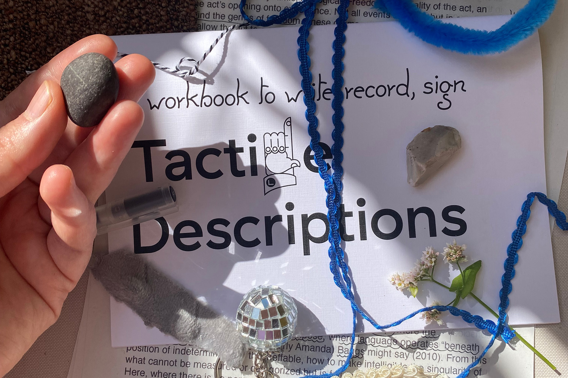 Various small objects, each with a different texture, are scattered across a brochure titled “a workbook to write, record, sign Tactile Descriptions.” From the left, a hand reaches into the frame, its fingers holding a small, cool stone. Lying on the brochure are textile elements such as delicate ribbons and fabric scraps, a flower with its stem, and a keychain featuring a glittering mini disco ball. The brochure itself rests on a printed text where terms like “nonlinear,” “perception,” and “force” are visible. Summer light and soft shadows create a playful atmosphere.