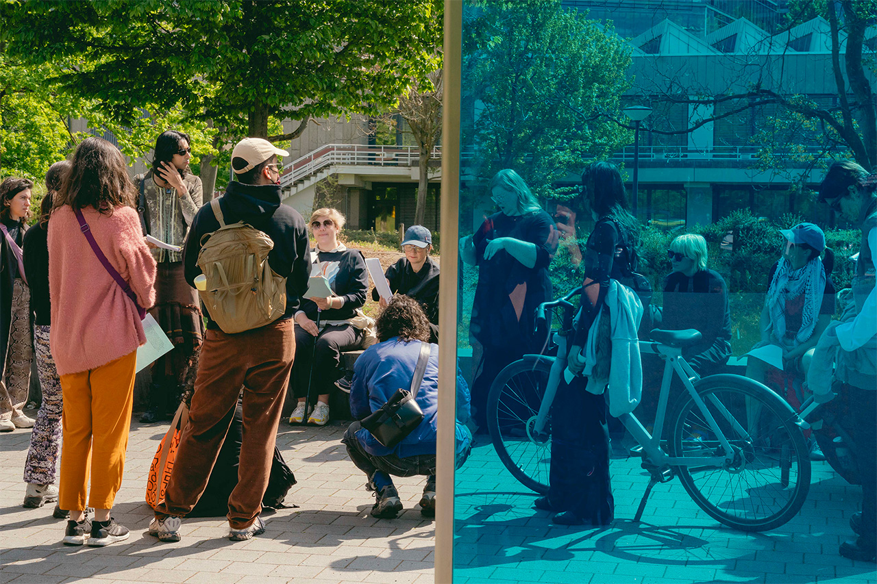 A group of people gathers in a half-circle at the T4 memorial with some of them standing, some sitting,  one kneeling, and one leaning against a bicycle. Half of the photo has been taken through the blue surface of a glass panel.